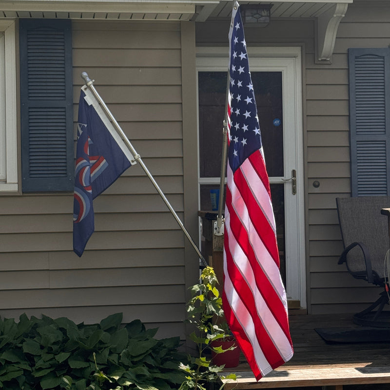 An American flag mounted on a pole outside a house, with a second dark blue flag hanging to the left, in front of a beige house with shutters and a porch.
